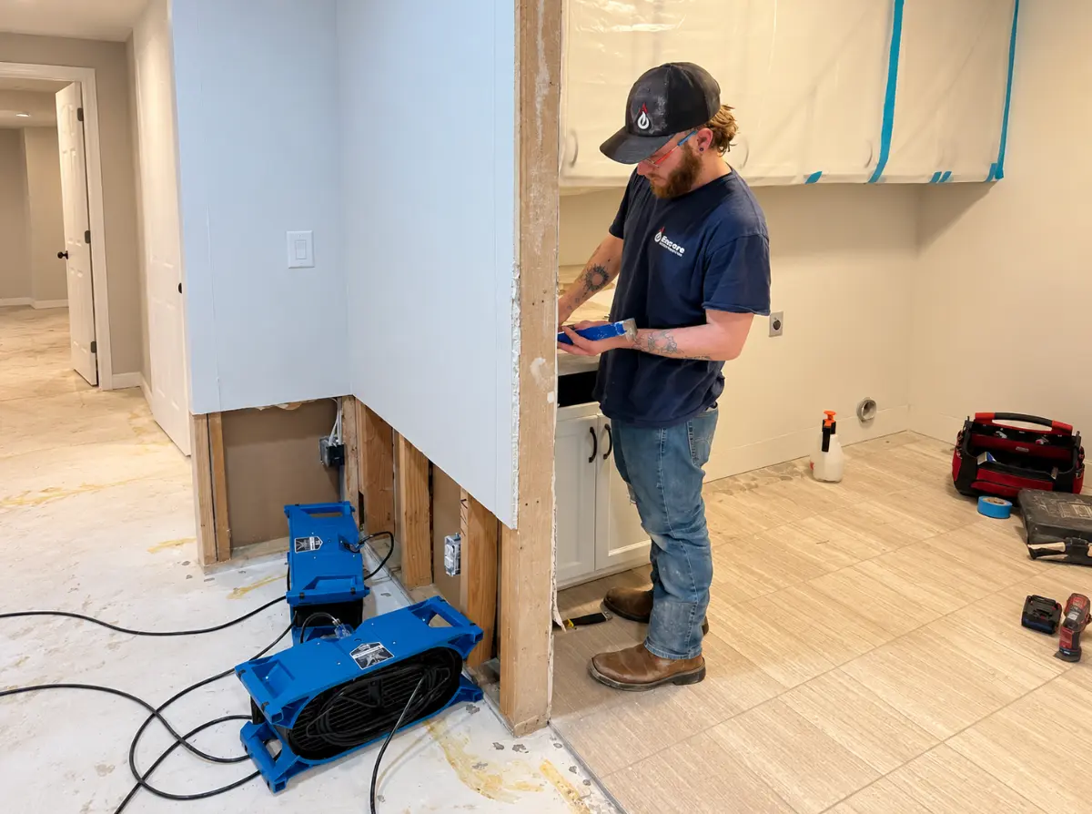Restoration technician working beside air movers in a partially opened residential wall.